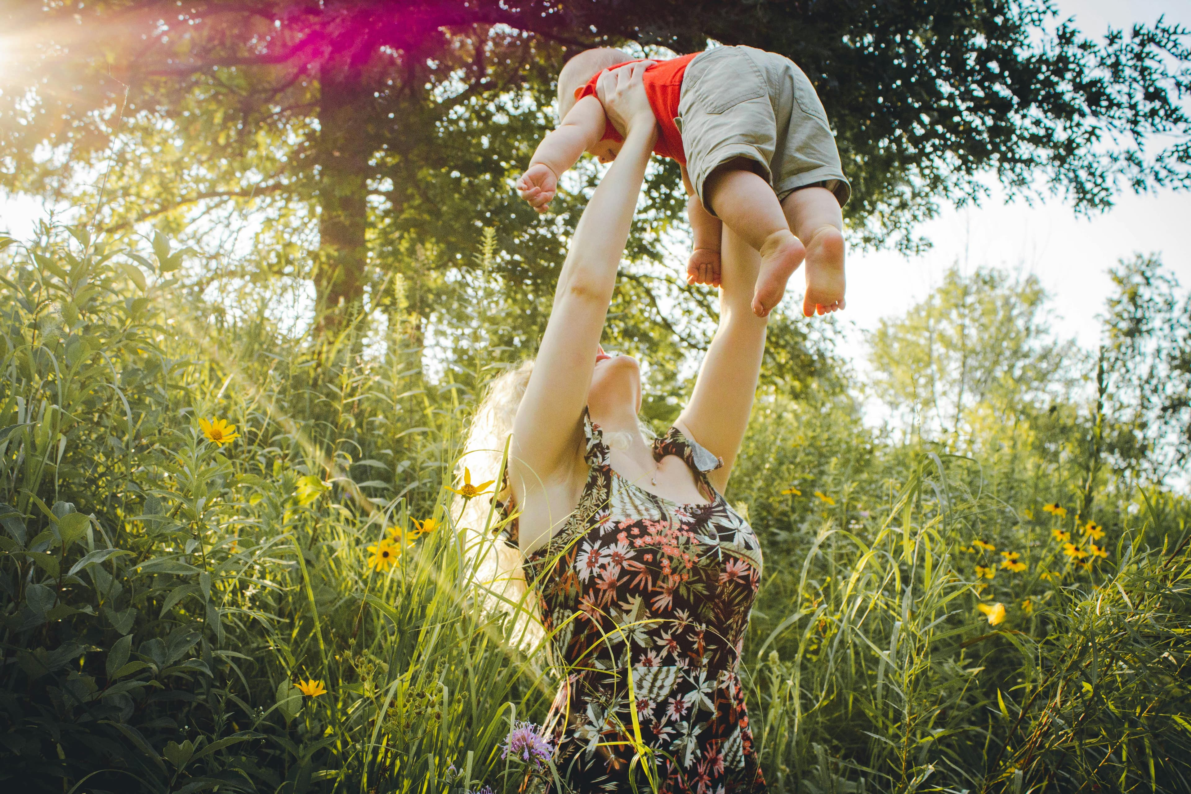 Mother lifting her baby into the air in a sunny meadow surrounded by tall grass and wildflowers.