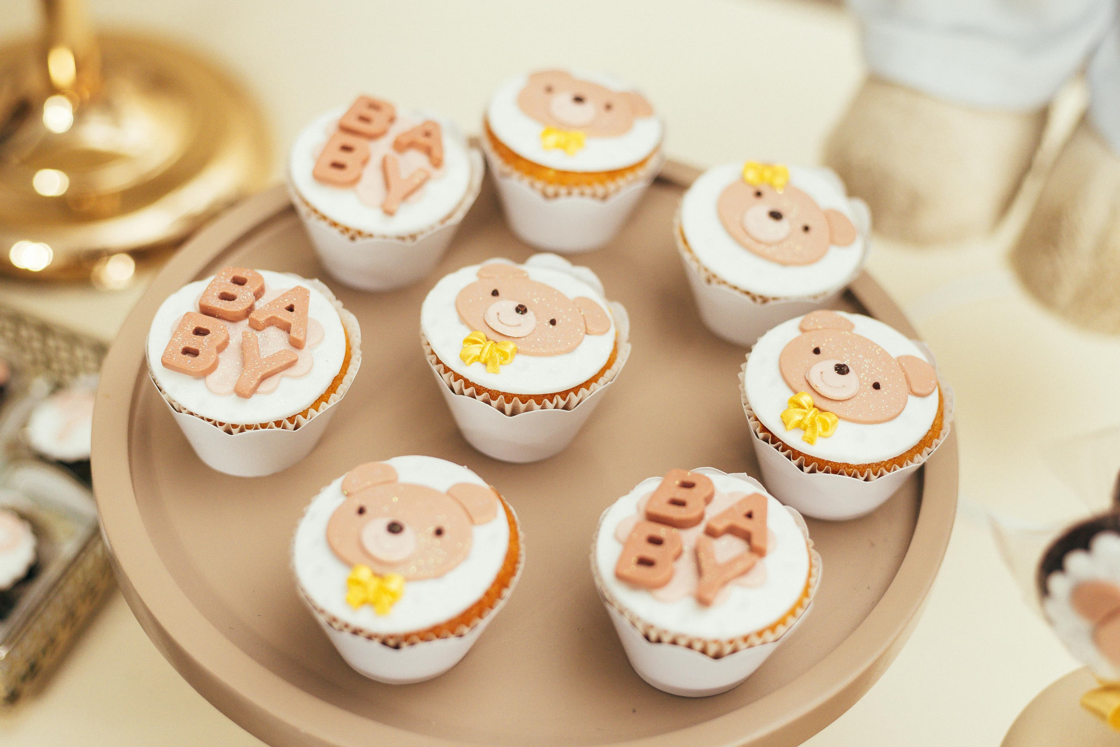 Plate of decorated cupcakes with teddy bear faces and ‘BABY’ lettering, arranged on a tray at a baby shower.