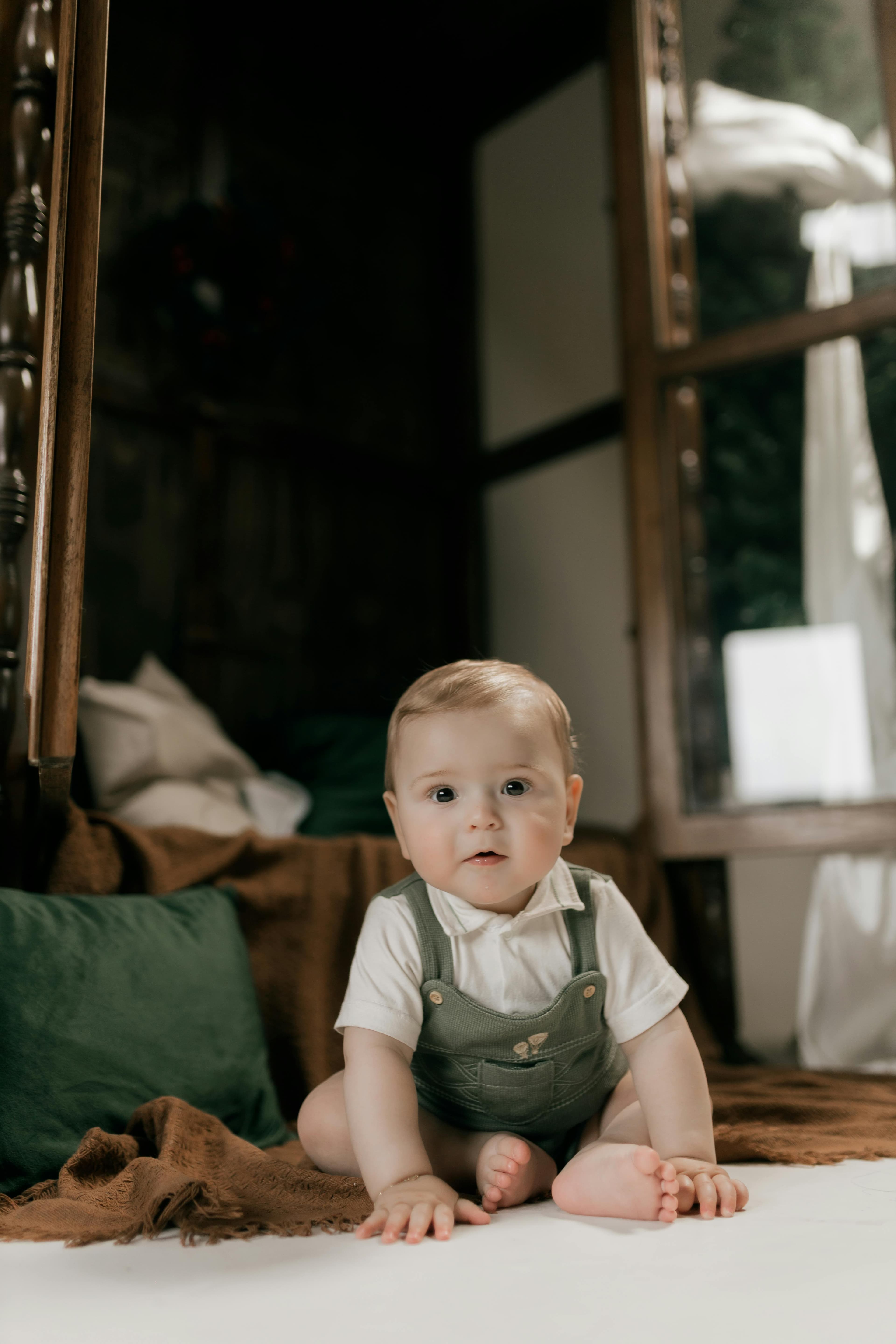 A baby sitting on the floor, wearing a white shirt and green overalls, indoors near a bed and a window with soft natural light.