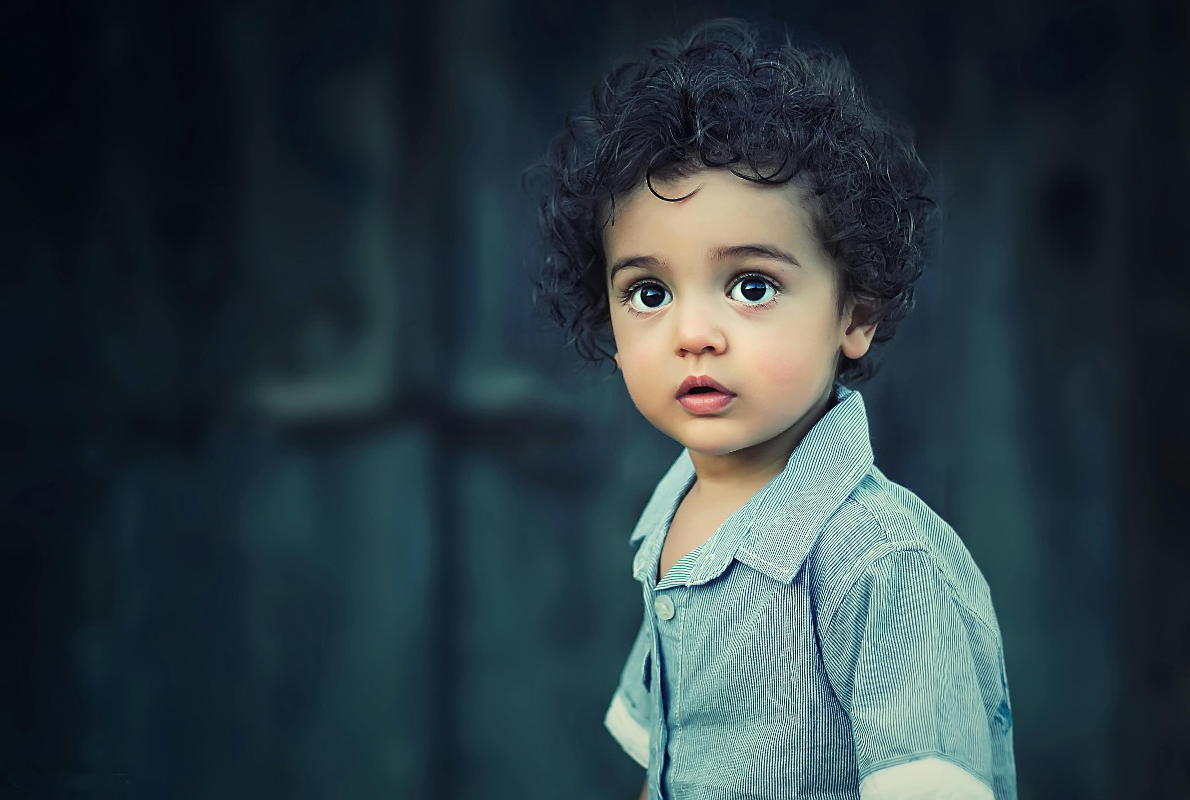 A young child with curly hair and large expressive eyes, wearing a light blue button-up shirt, looking slightly to the side against a softly blurred dark background.