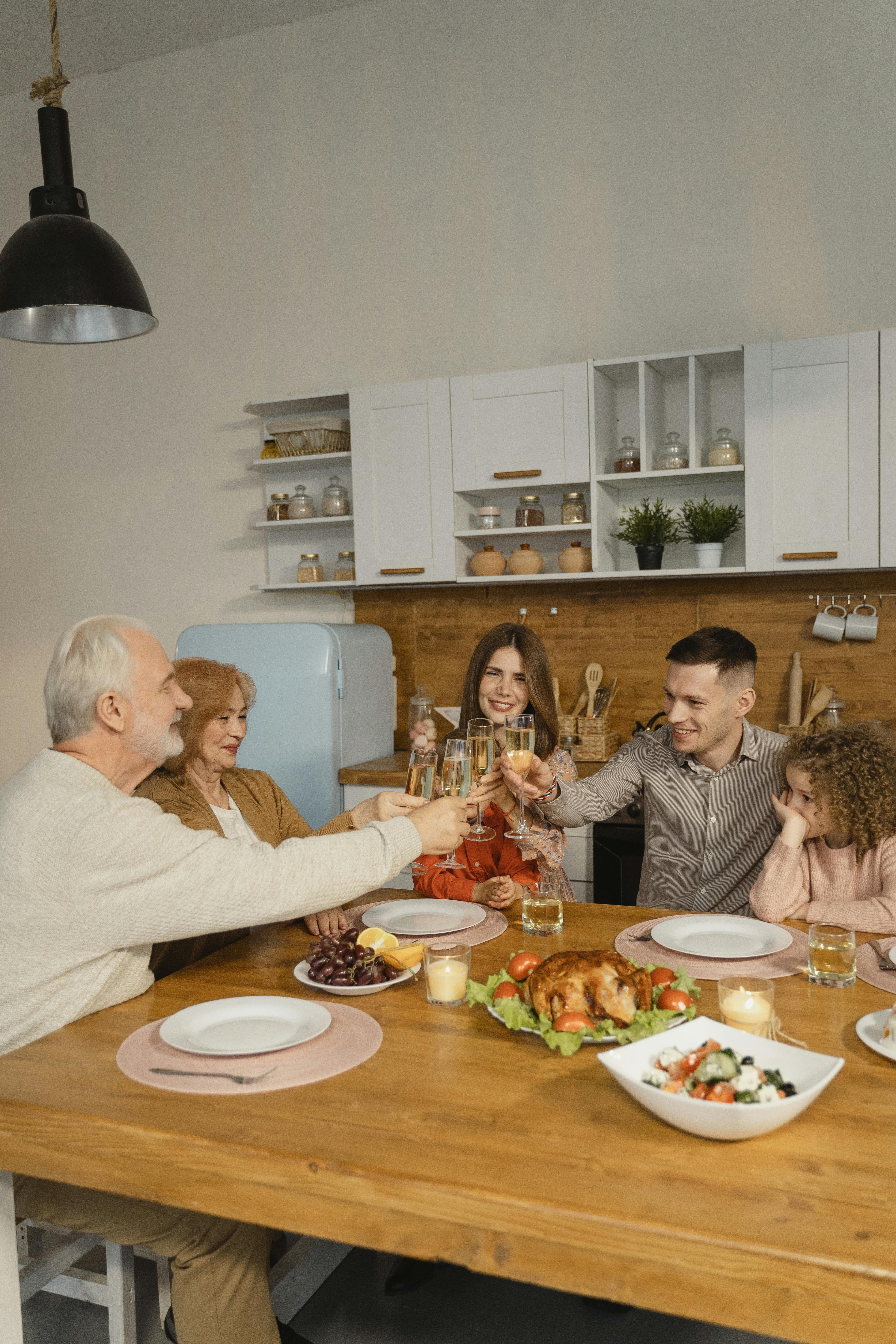 Family gathered around a dining table in a kitchen, toasting with glasses while sharing a meal that includes roasted chicken and side dishes.